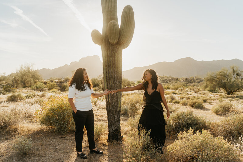 A couple reaching out and holding hands in front of a cactus 