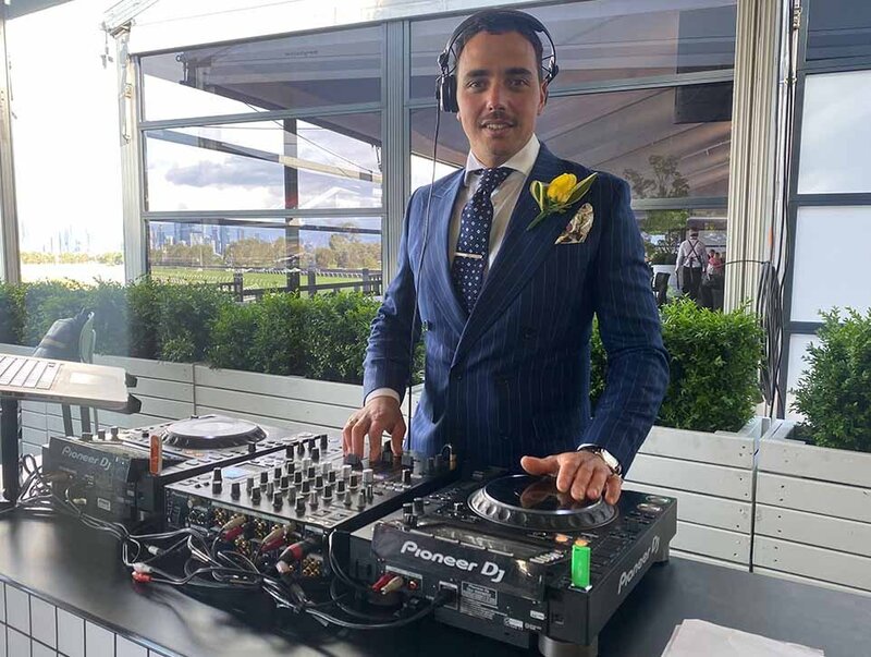 Male DJ in a navy pinstripe suit with a yellow boutonnière mixing music on Pioneer DJ decks at the Melbourne Cup Carnival