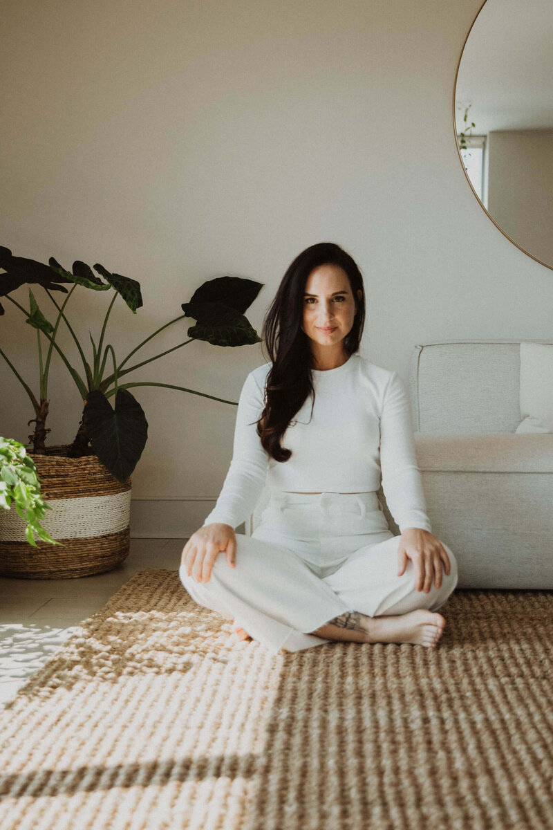 Ayurvedic skin specialist, Jen, sitting on a jute rug with her legs crossed wearing a white top and pants