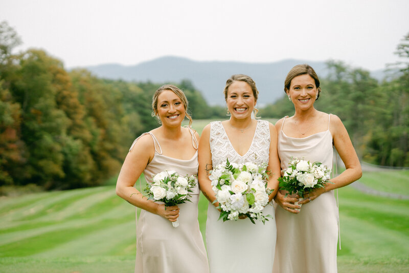 Three women in light dresses stand smiling outdoors, holding bouquets of white flowers. A grassy landscape with trees and mountains is visible in the background, under an overcast sky.