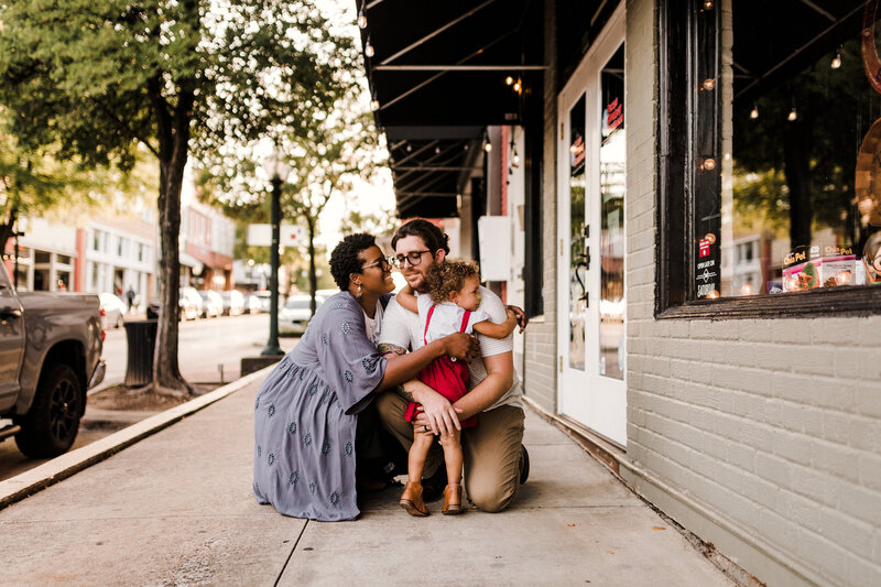 Photograph of a family in the street, the woman is wearing a light blue cardigan and is looking at the child who is on his back hugging the man who is kneeling in light brown trousers with a white T-shirt