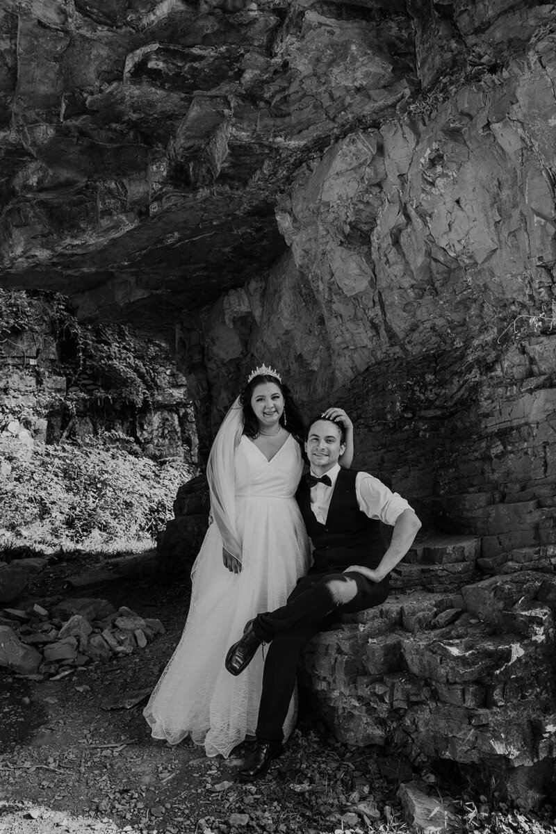 A groom sits on a chimney rock while his bride snuggles in, and caresses his head. She is wearing a tiara and veil, and he is wearing a vest and bowtie