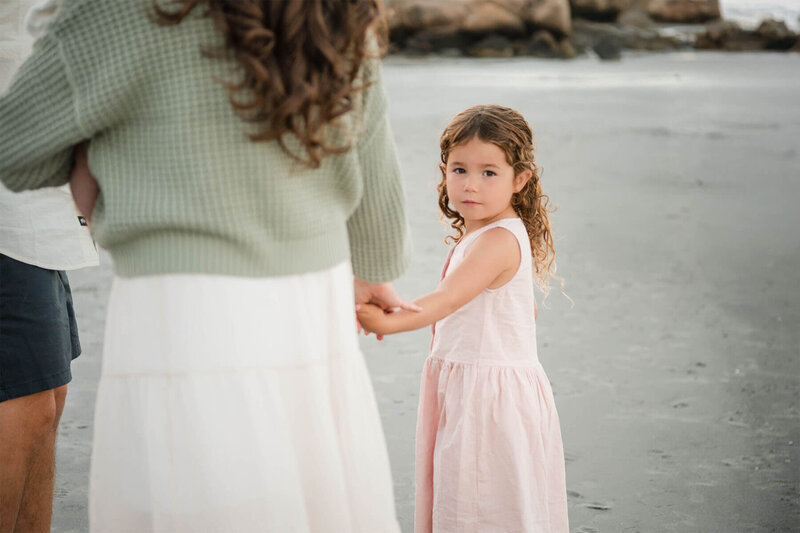 A little girl holds her mom's hand and looks back at the camera as she walks on the sand at Wingaersheek Beach.