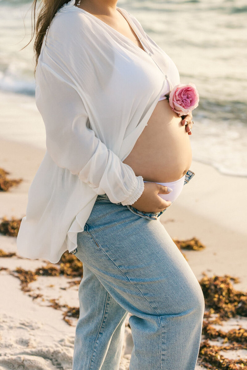 Mom holding her baby bump and a pink rose above her belly at a beach in Miami, FL.
