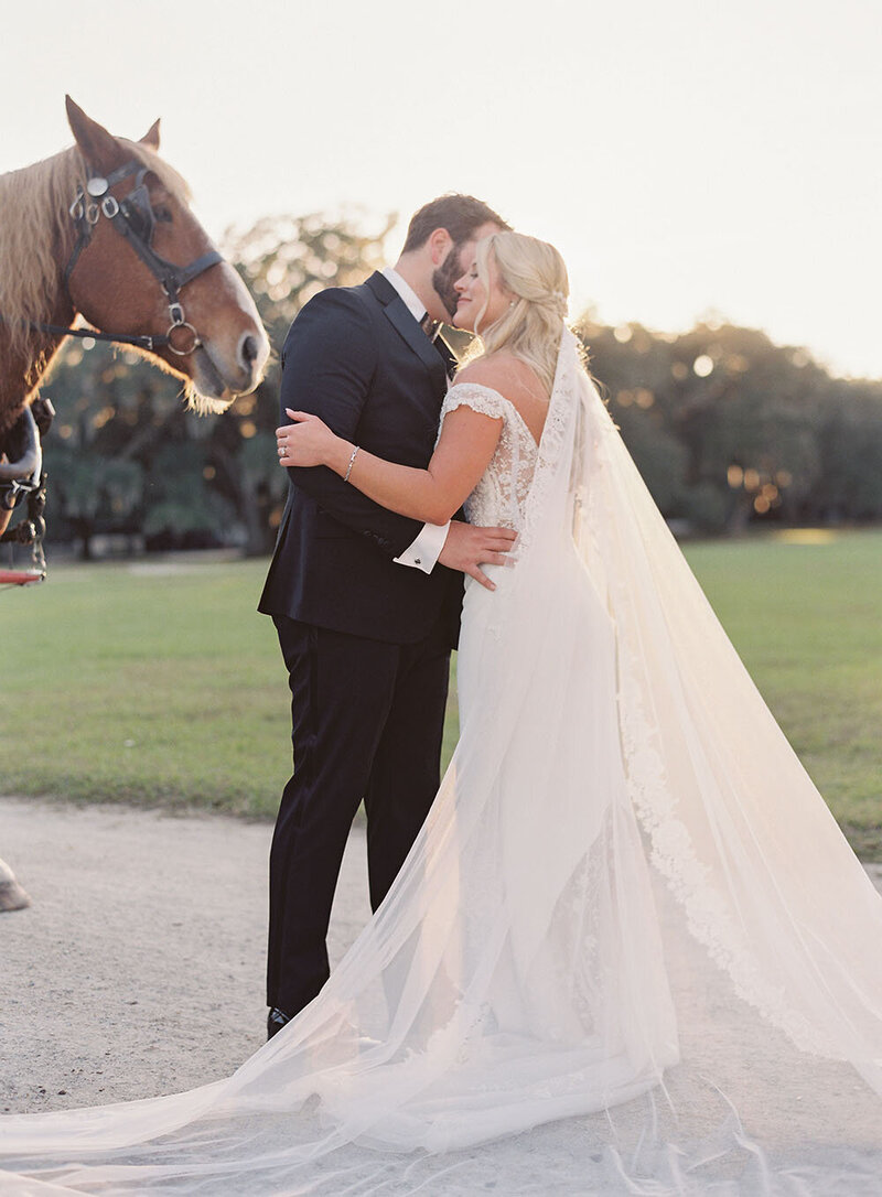 Off the shoulder wedding dress with an embellihsed bodice and dramatic veil hugging her groom in a black tux next to their horse and carriage