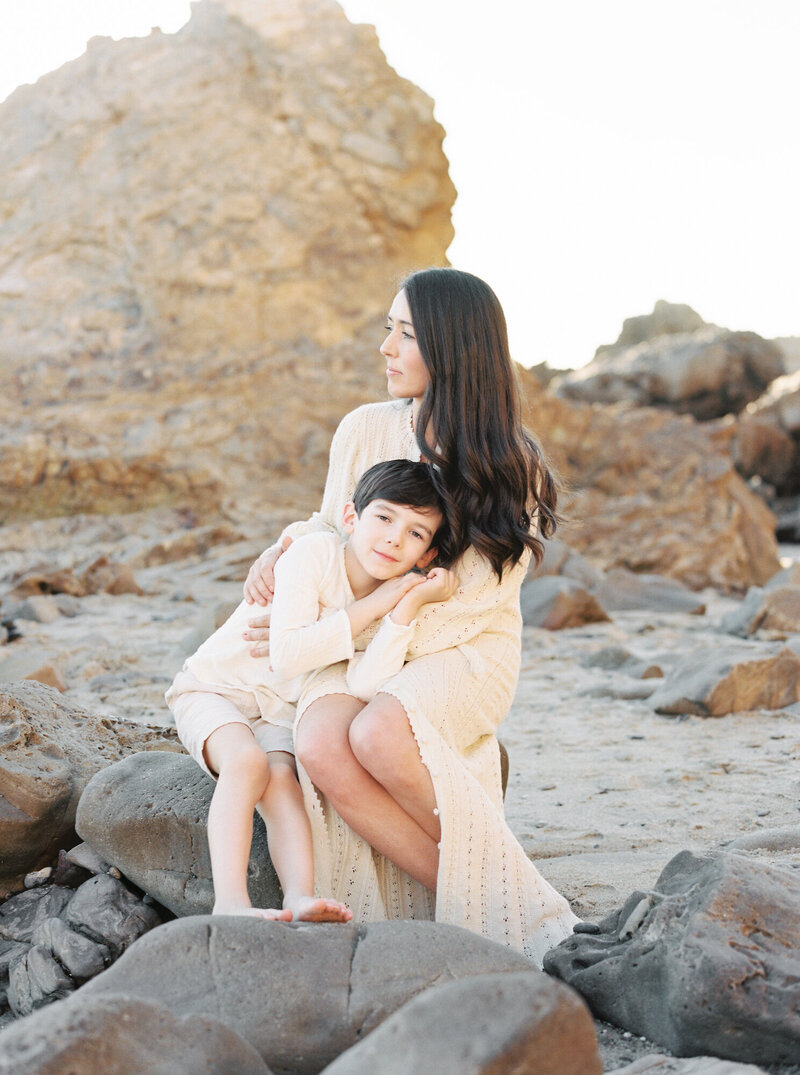 portrait of Seattle Newborn and Maternity Photographer Brittany Carvalho with her son on a rock at beach in Orange County wearing cream outfits 