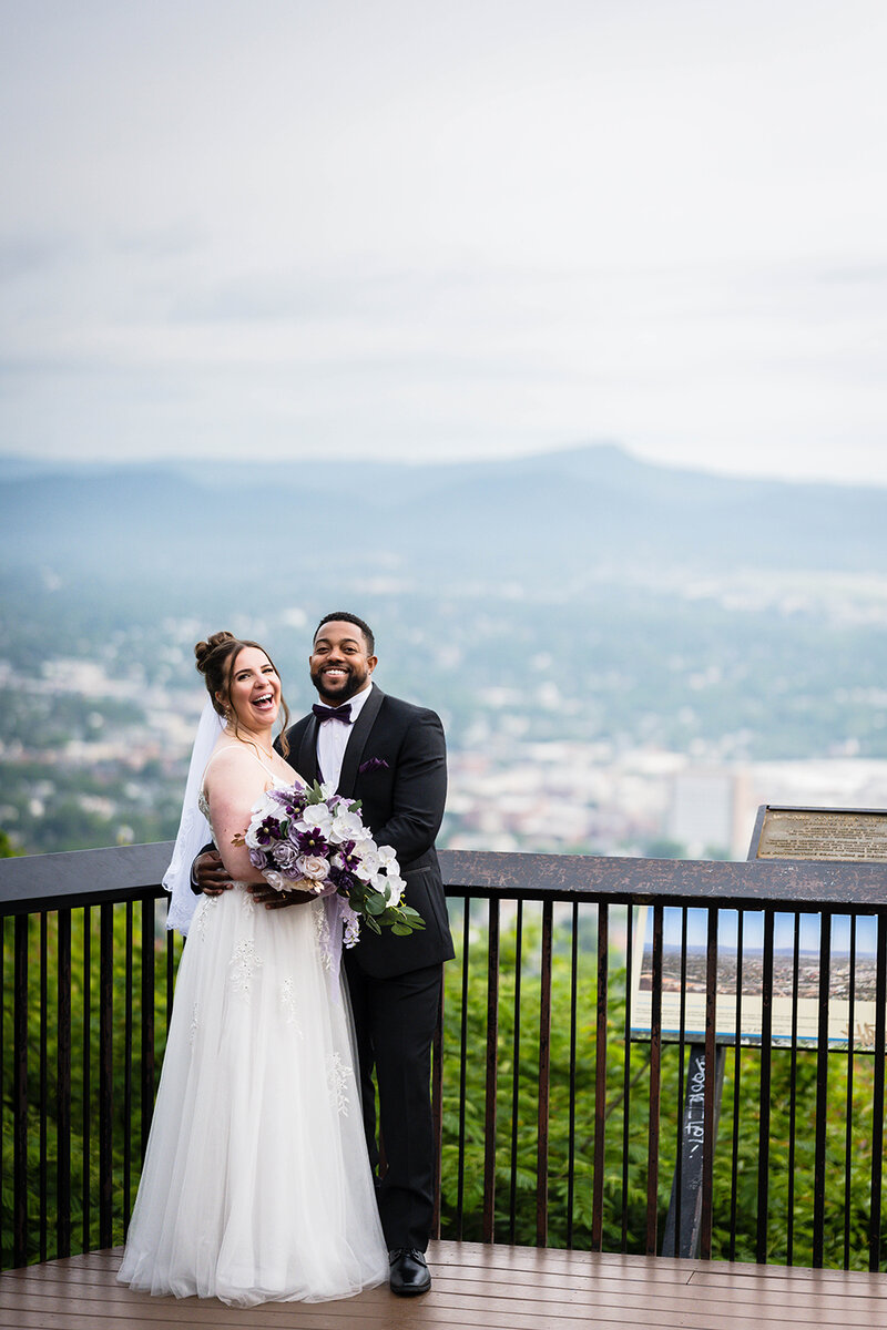 A couple smiles widely as they pose together after their wedding ceremony at the Mill Mountain Star Overlook in Roanoke, Virginia. The background is of the Blue Ridge Mountains and the city of Roanoke.