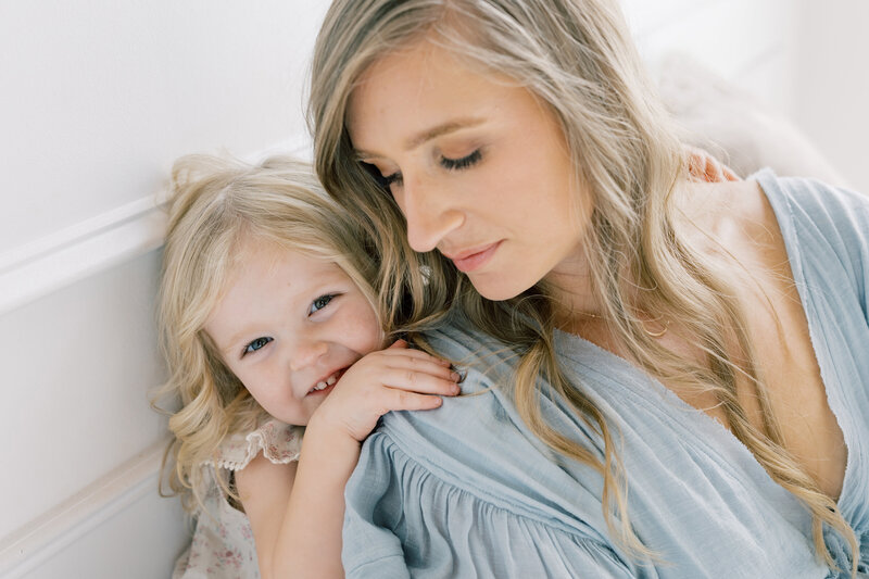 Mother hugging daughter closely, both smiling softly in a natural light setting.