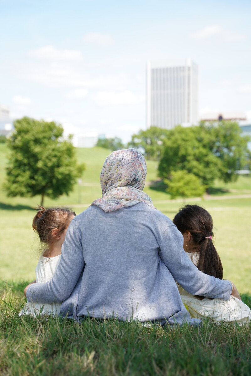Fitness hijabi coach Hanan from behind with her two toddler daughters, one on each side, sitting on the grass in a park