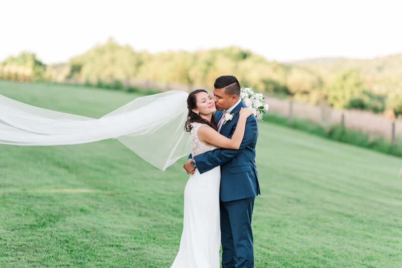 Groom wraps his arms around his new bride, her long cathedral veil swept by the wind behind them. The background shows views of rolling Virginia mountains, lit up in orange and yellow by the sunset.
