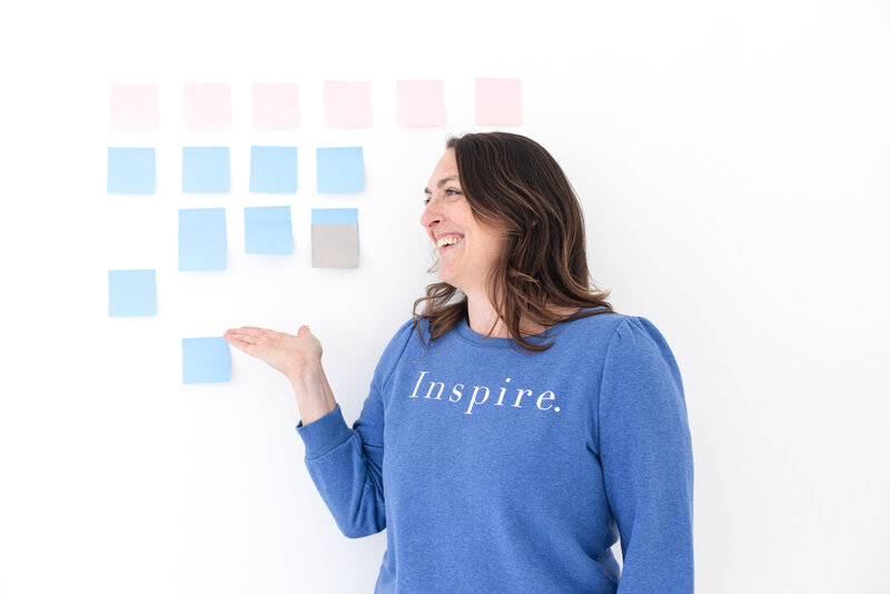 the creator of courses for business owners is pointing to the light blue sticky notes on the white wall with her right hand. the social media strategist is smiling and wears a blue coat with a white inspirational word