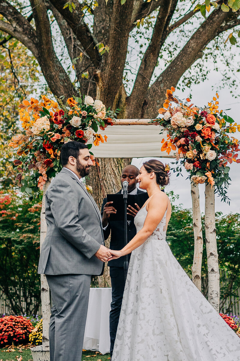 A bride a groom and their officiant exchange vows at Feast at Round Hill in New York's Hudson Valley before a birch chuppah decorated with large autumn flower arrangements