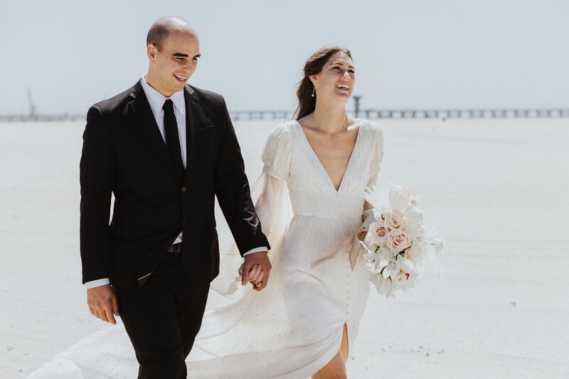 Wedding Photographer, bride in her dress and groom in his tux walk hand-in-hand down the beach