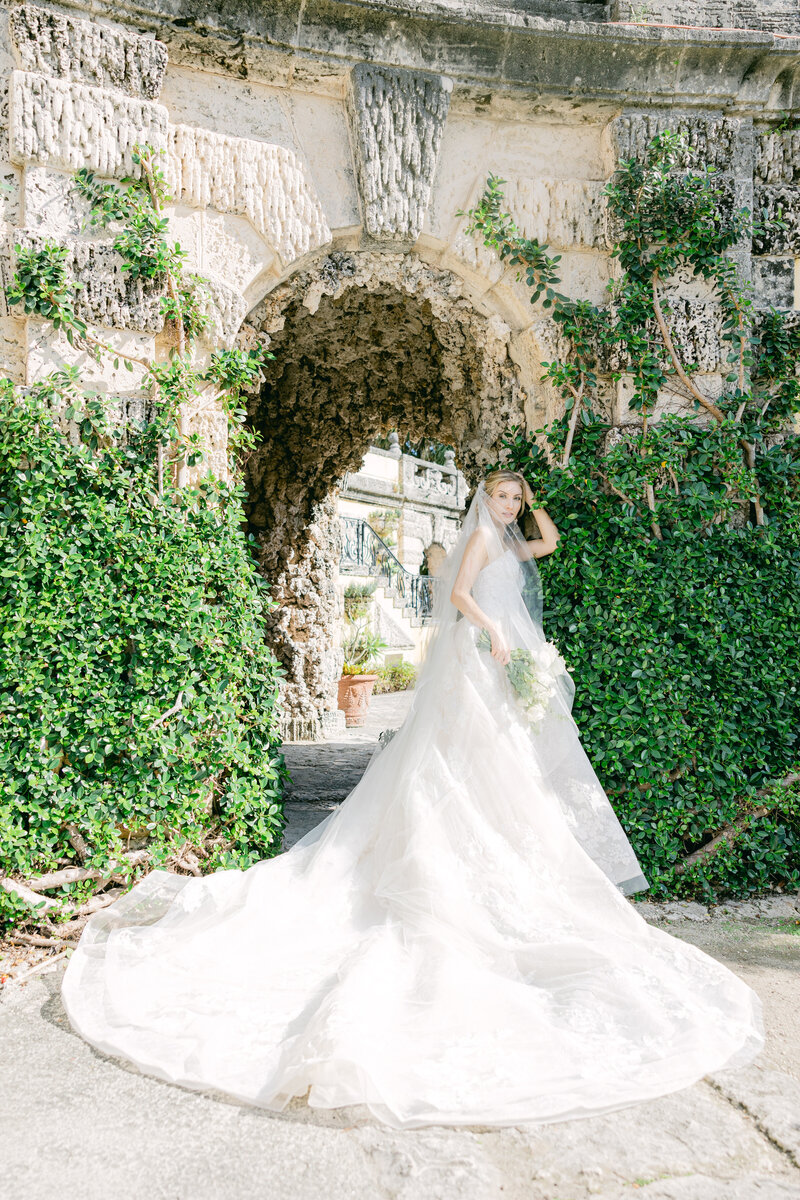 A breathtaking shot of a bride walking through a lush, ivy-covered archway at the Vizcaya Museum in Miami. She wears a magnificent gown with a long train, surrounded by the historic and romantic architecture of the museum. The image captures the elegance and timeless beauty of this iconic venue, known for hosting unforgettable weddings. The bride gracefully holds her bouquet as the natural light illuminates her veil and the intricate details of her dress.