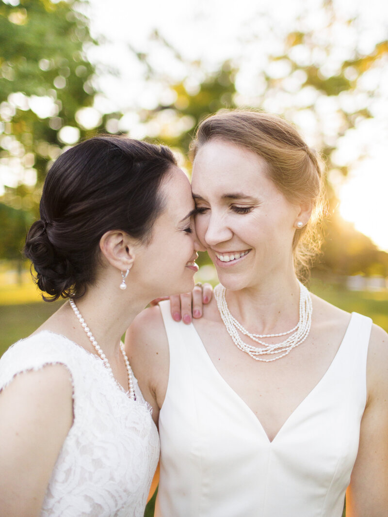 Two women in wedding dresses embrace and smile at each other outdoors. They wear pearl jewelry, and sunlight filters through trees in the background, creating a warm, intimate atmosphere.