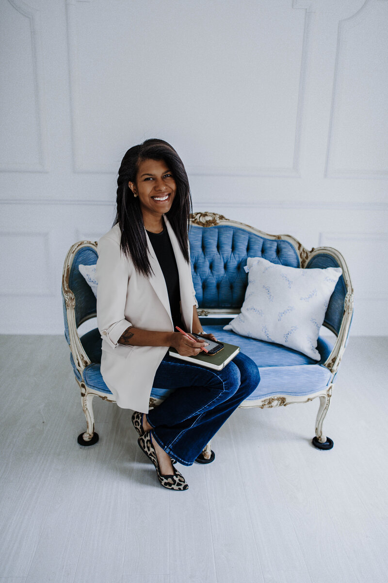 Black Female Photographer sitting on Blue couch in studio. She is holding a notebook and phone and smiling at the camera.
