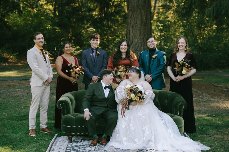 Brides smiling at each other on a sofa at outdoor wedding with family behind them. 
