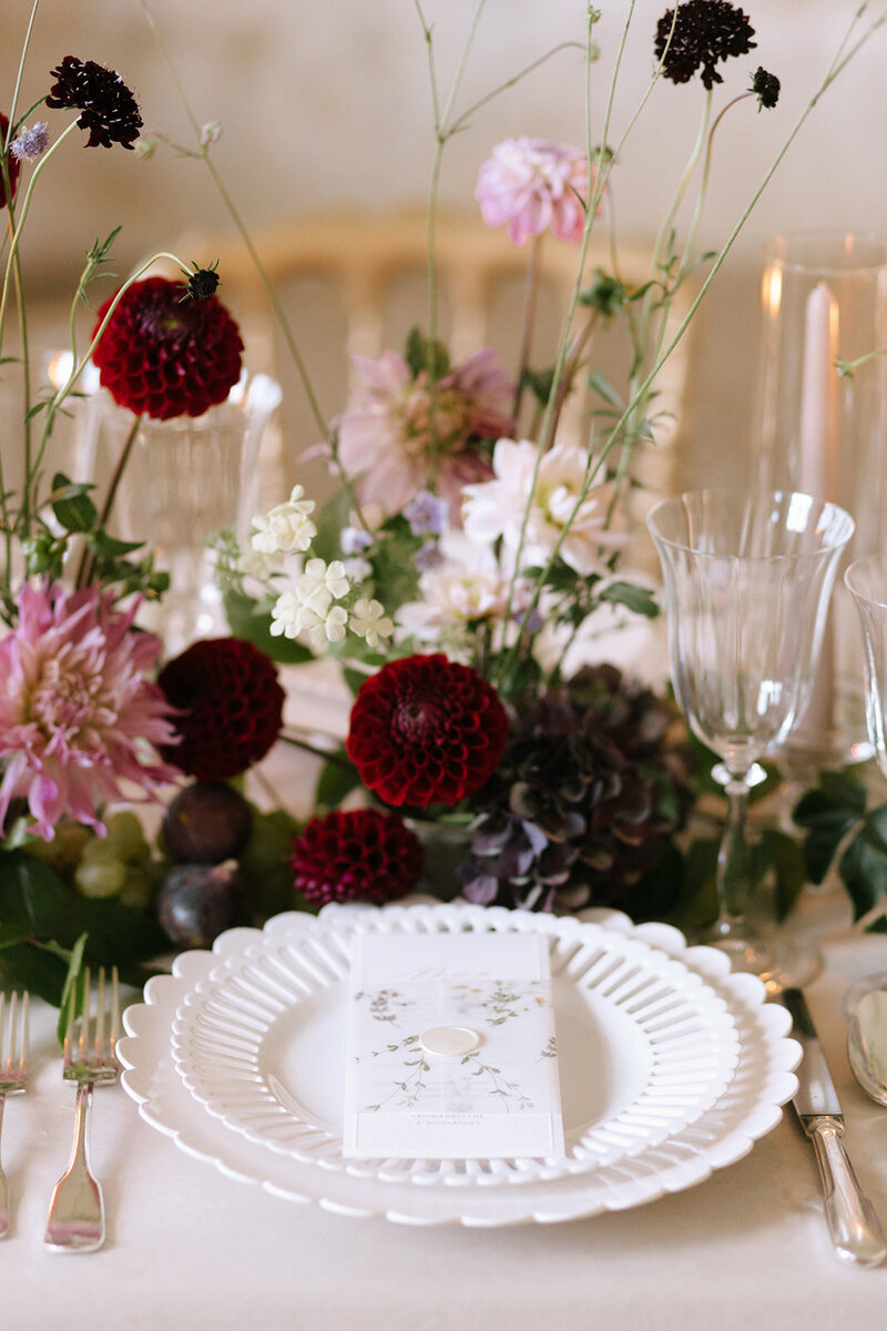 A romantic wedding tablescape styled with burgundy and blush flowers, delicate glassware, and a white scalloped charger plate with a floral-printed menu card,