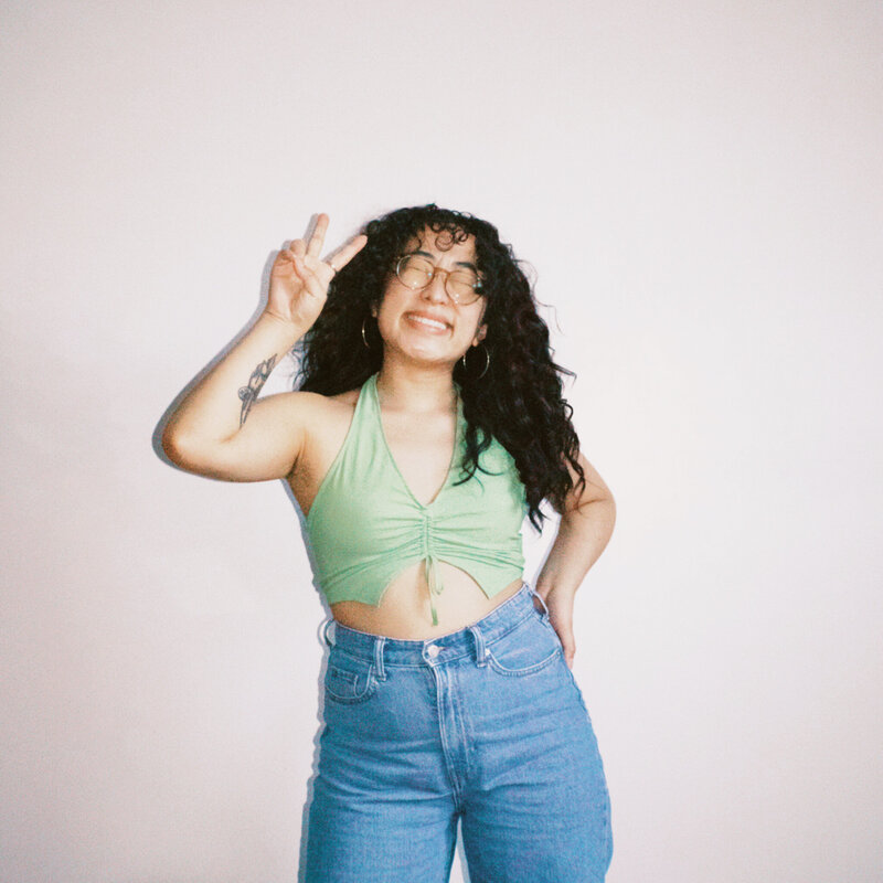 Asian woman smiling with curly hair and light green crop halter top and blue jeans, throwing up a peace sign