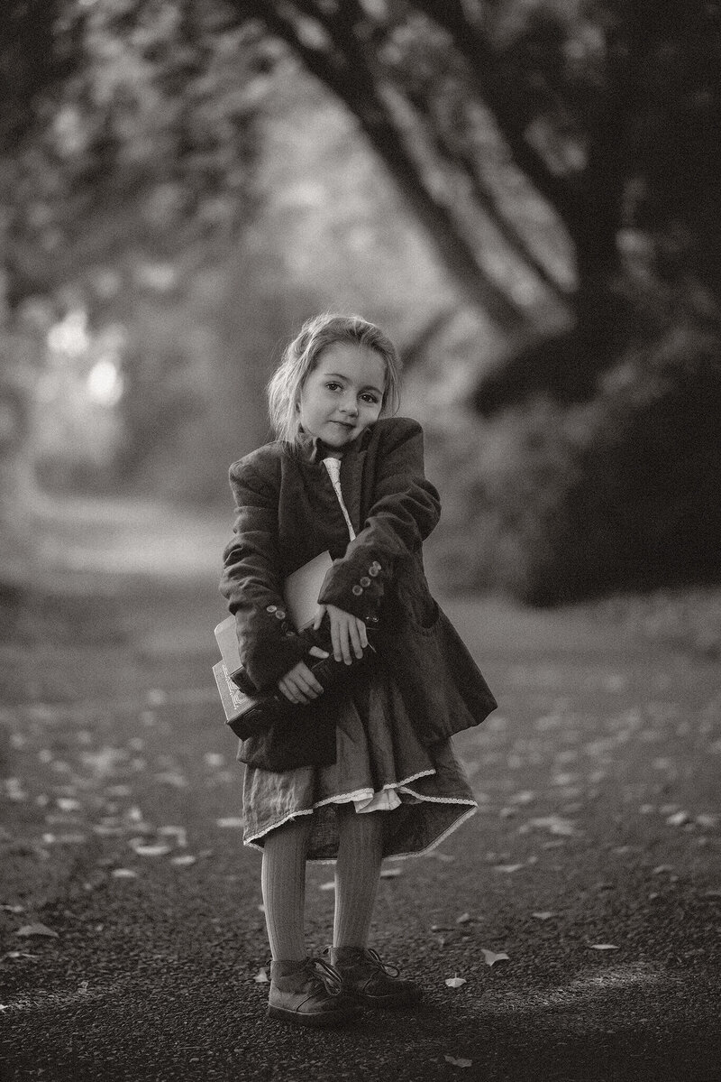 An up close black and white portrait of a little girl smilling as she hold a stack of old books in her hands. She is wearing a coat with a dress underneath