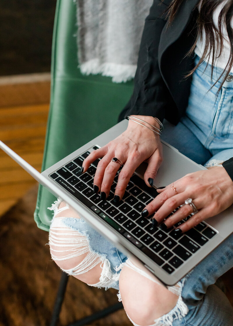 Close-up of Allie Clark’s hands typing on a MacBook Pro, seated on a green leather chair, visually capturing the detail-focused web design work that powers Allie Clark Designs’ elevated custom websites.