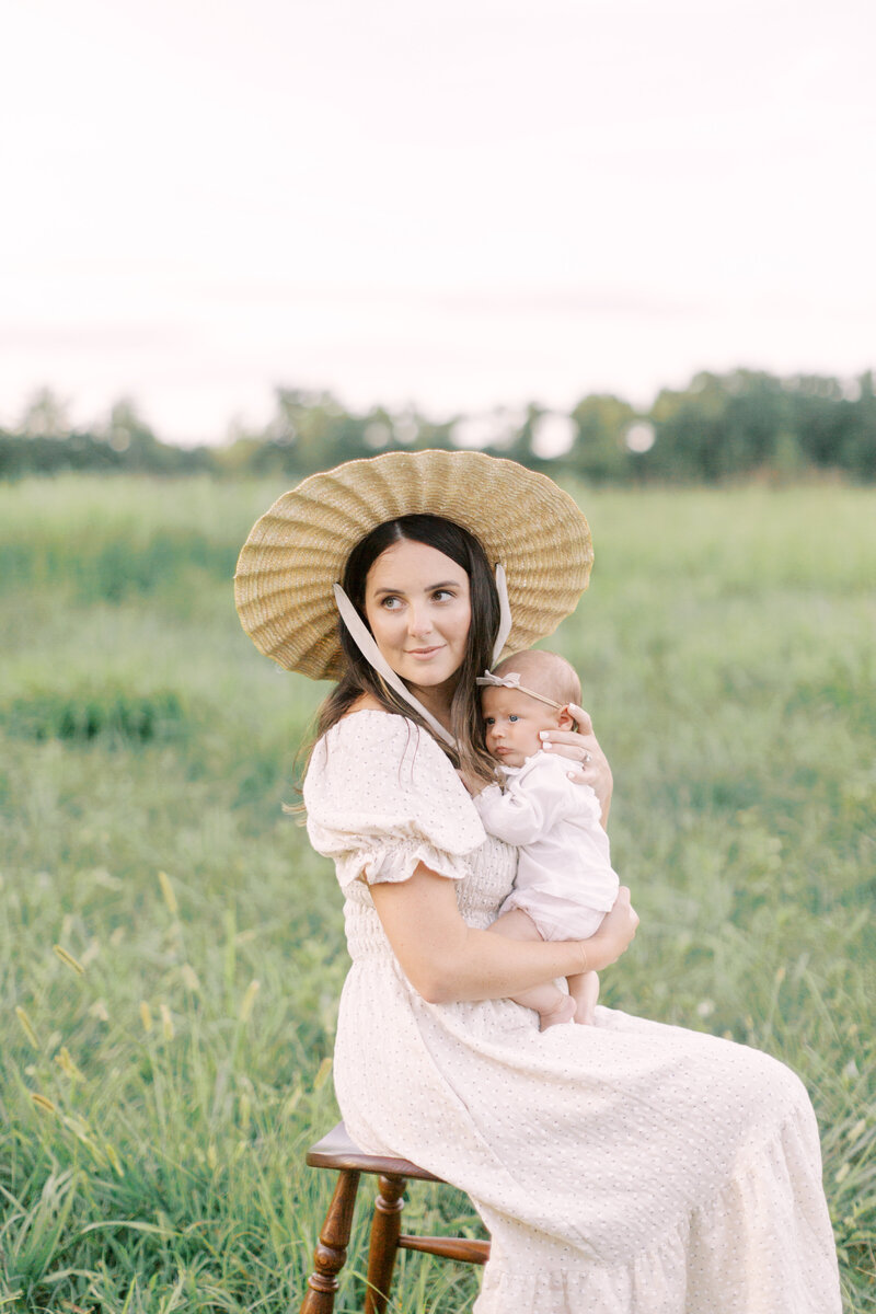 Mother wearing a hat sitting on a stool in a field holding her newborn
