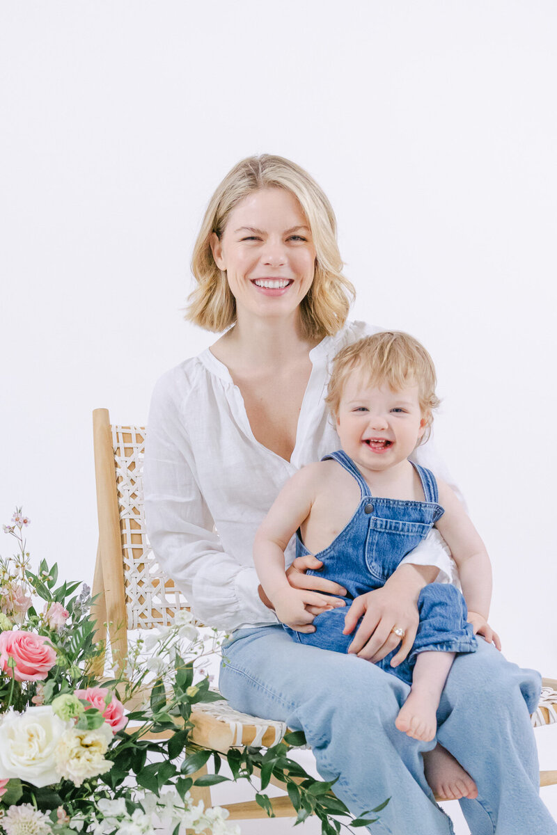 A joyful mother with short blonde hair and a white blouse sits on a woven chair holding her smiling toddler, who wears denim overalls. Delicate pink roses and greenery frame the bottom of the image. Captured in a bright white studio by Nicole Neira Photography, this portrait radiates warmth, love, and the beauty of everyday motherhood.