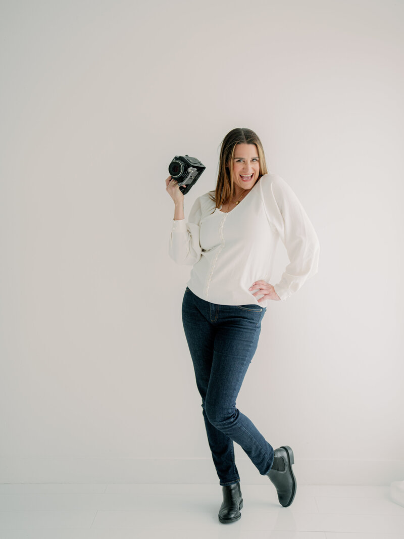 Courtney Bowlden smiling at camera in front of a white wall holding a camera in one hand and the other on her hip