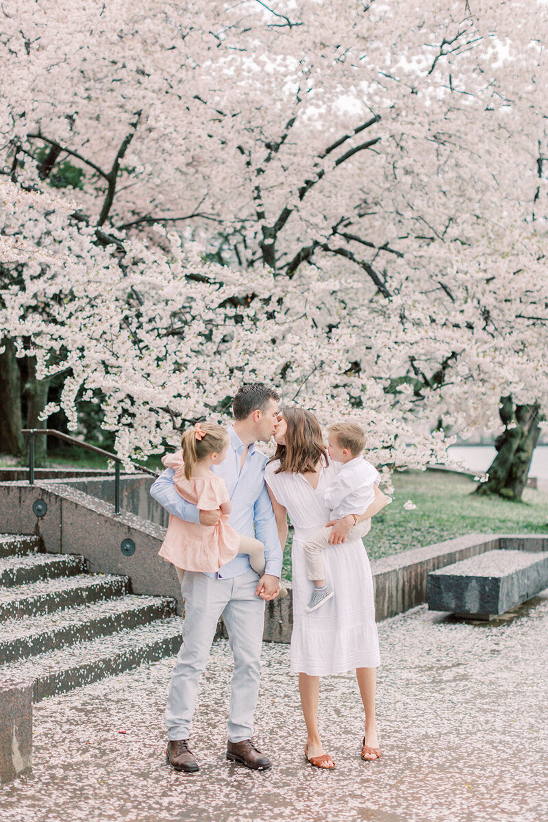 A mother and father stop to kiss while holding their two young children while standing in the cherry blossoms at the Tidal Basin, photographed by Family Photographer Northern Virginia Marie Elizabeth Photography