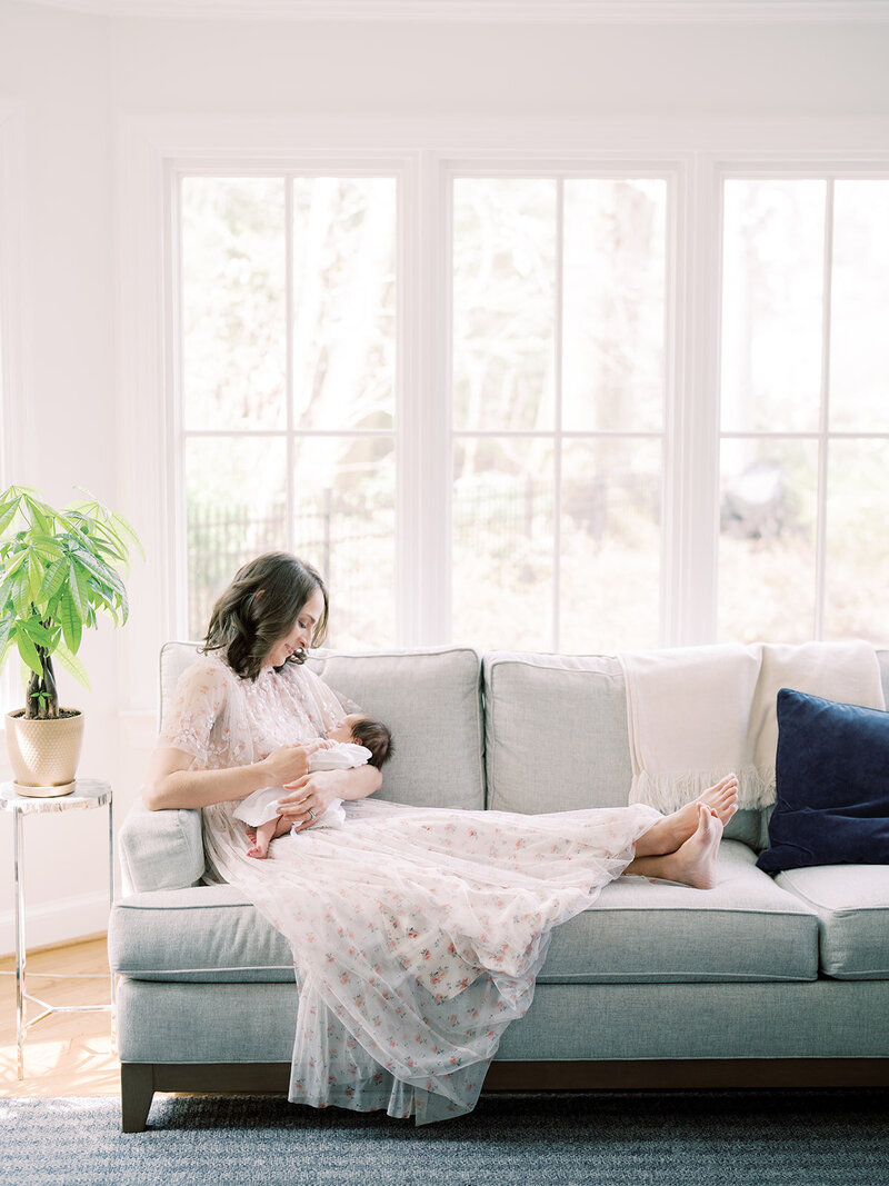 Mother sits on the edge of a couch with her feet up on the couch looking down at her newborn baby girl.