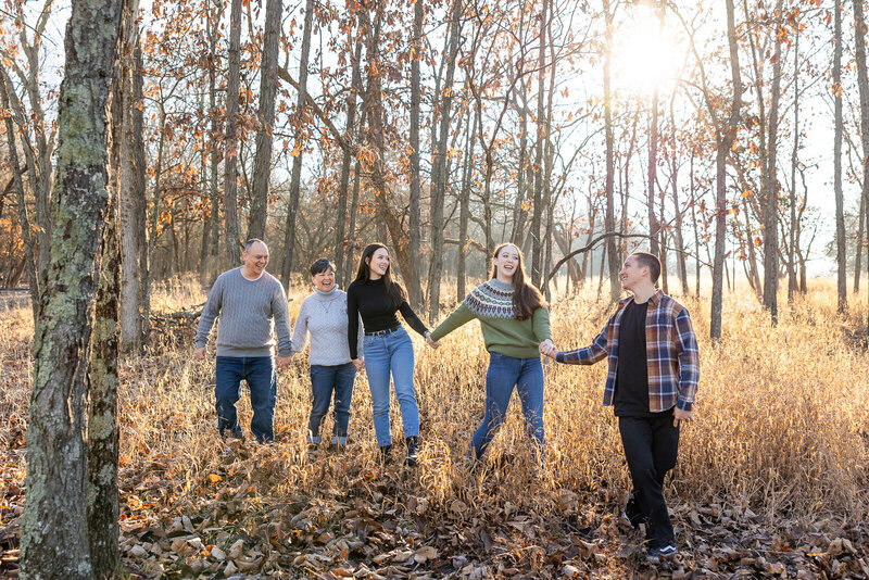 Family of five with adult children and two parents all holding hands walking in field of tall grass. 