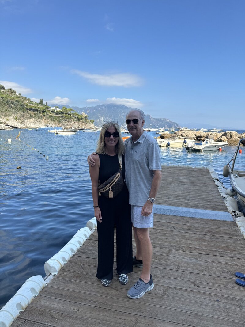 Couple standing on wooden dock on Amalfi Coast with mountains in background. 