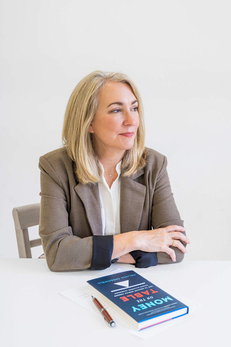 Melissa Greenwell, sitting at a desk with her book, Money on the Table