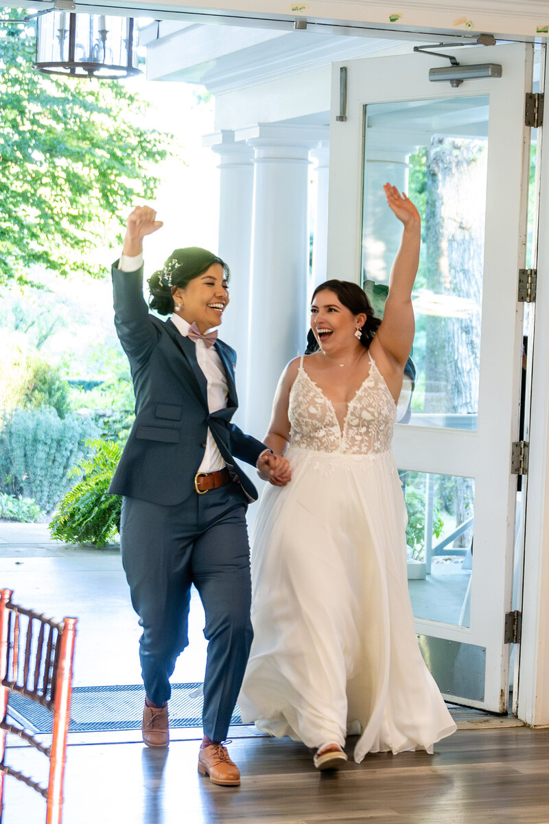 laotian bride in blue suit and a curvy white bride in a spaghetti-strap gown holding hands and smiling with hands raised in the air walking into a wedding reception 