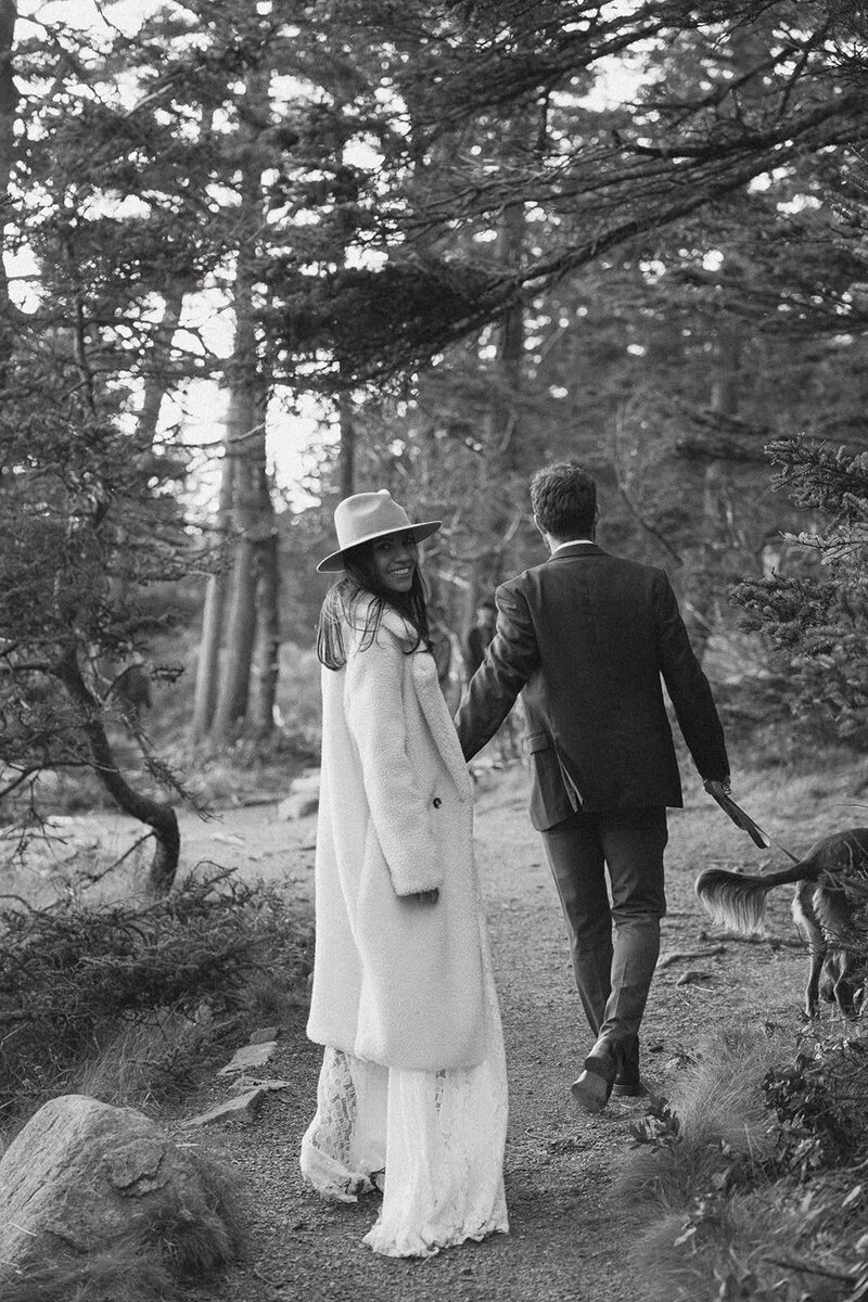 Black and white photo of bride holding grooms hand and looking back at the camera while they are walking their dog in Acadia on their wedding day