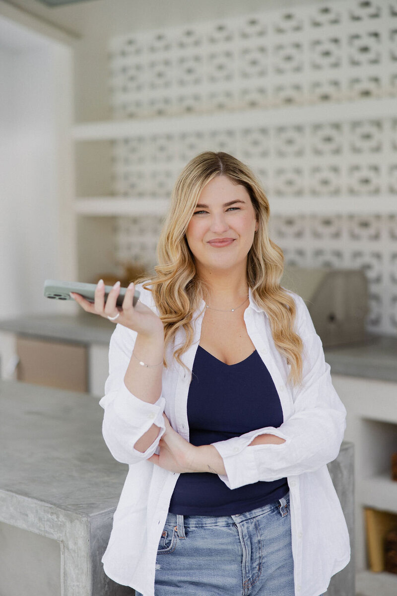 Brogan leaning against a modern outdoor patio area holding up a phone, wearing a loose white button up