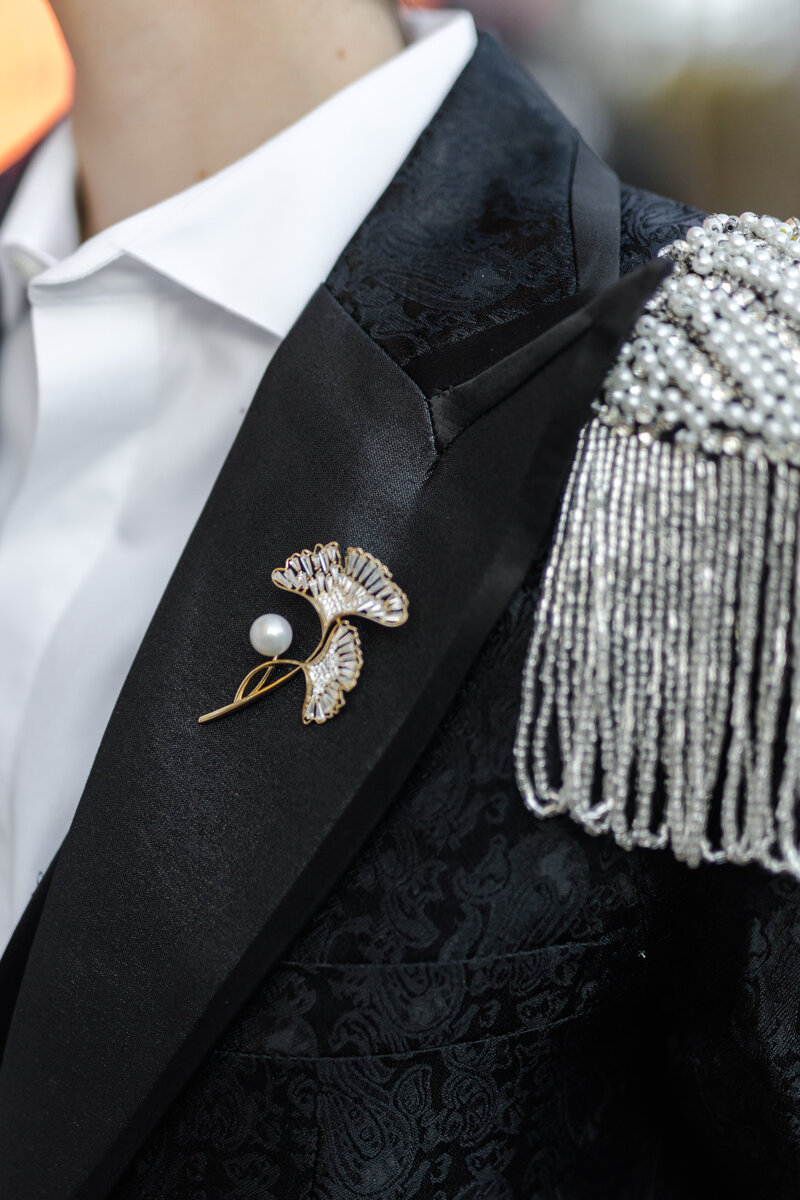 Closeup of the nonbinary newlywed in their black shimmer suit with pearl sequin lapels and a gold, peal, crystal ginkgo leaf pin on their satin black lapel
