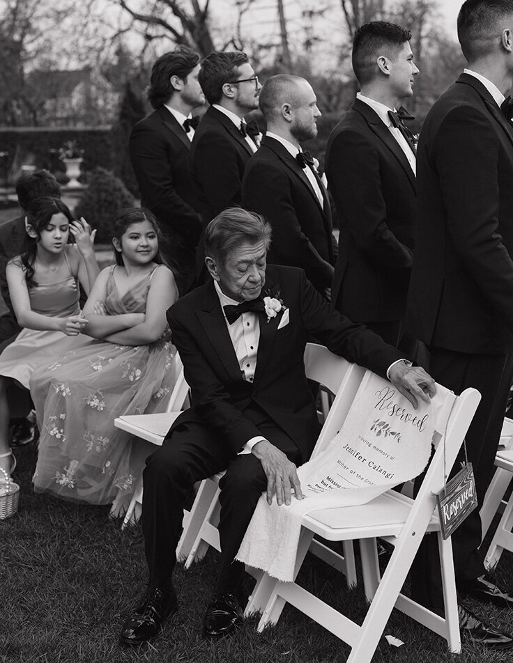 A black and white photo of an elderly man in a tuxedo sitting in a row of white chairs at an outdoor event. He glances at a reserved sign on the chair beside him. Nearby, girls in formal dresses and a group of men in suits stand, creating a somber, formal atmosphere.