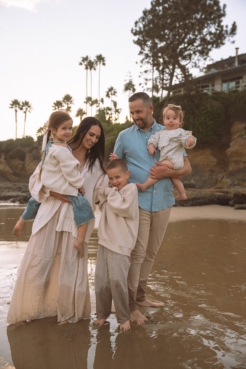 A family of five standing barefoot on the wet sand at Laguna Beach during golden hour, smiling together near the waterline with palm trees and coastal cliffs in the background.