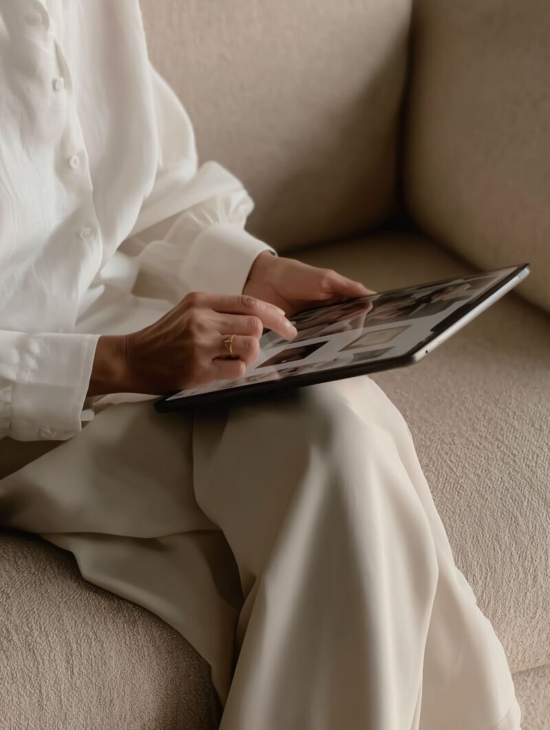 Close-up of person in cream blouse and trousers sitting on a beige sofa while browsing design inspiration on a tablet