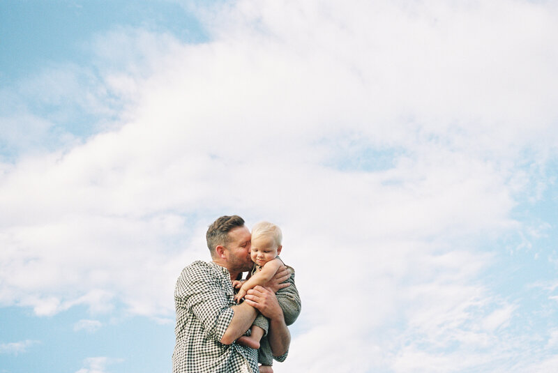 Photo from below of dad kissing baby son with blue sky and clouds behind them
