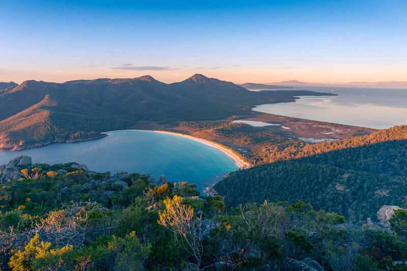 A scenic aerial view of a crescent-shaped bay with turquoise water surrounded by forested mountains and a sandy beach, taken during sunrise or sunset.