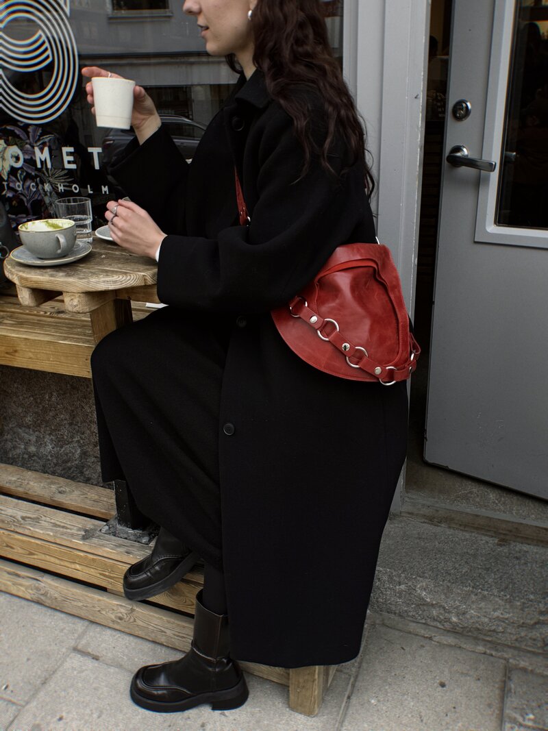 A writer is leaning over a coffee table, a grey mug in front of her, wearing a black trench coat, black combat boots, and a red shoulder bag