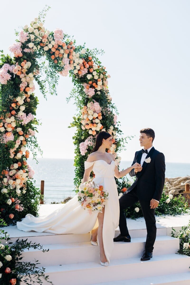 A couple under a colorful floral arch at a wedding ceremony by the sea. This wedding ceremony was design and planned by Thater Made, a luxury wedding an event producer based in San Diego, CA.