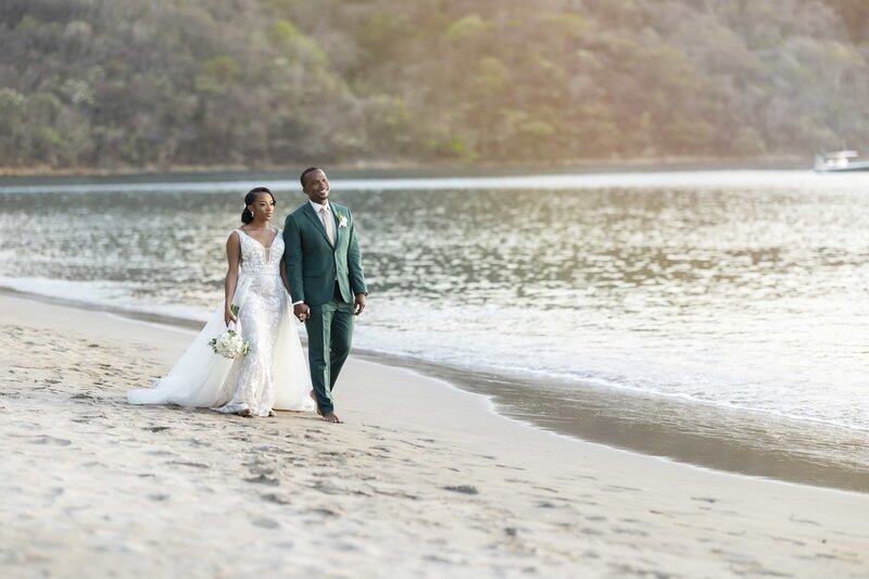 A bride and groom walking on the beach near a body of water.