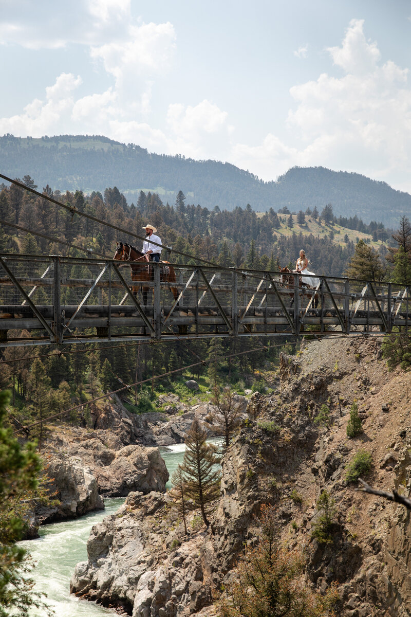 Bride in a flowing wedding dress and groom in a cowboy hat ride horses across a suspension bridge high above a rugged river canyon, surrounded by forested mountains.