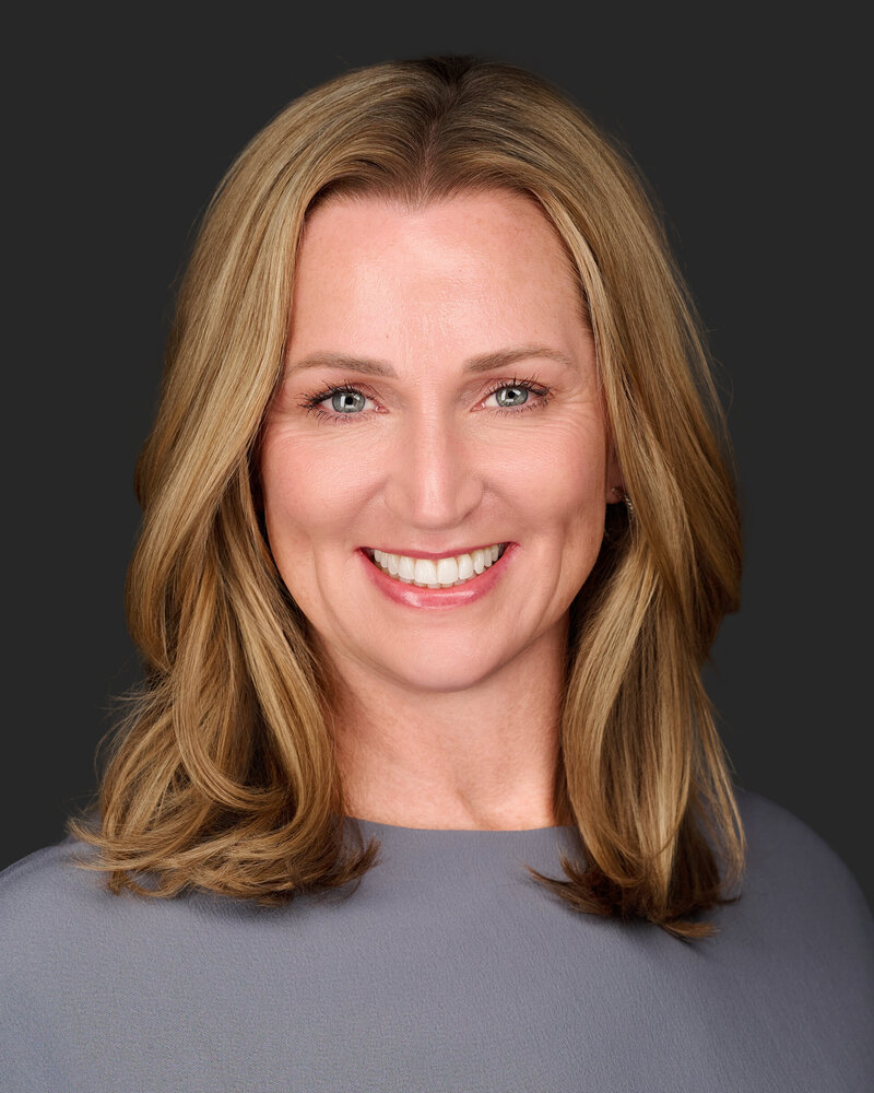Jenn Cloutier professional headshot with warm smile, shoulder-length blonde hair, wearing a gray top against dark background.