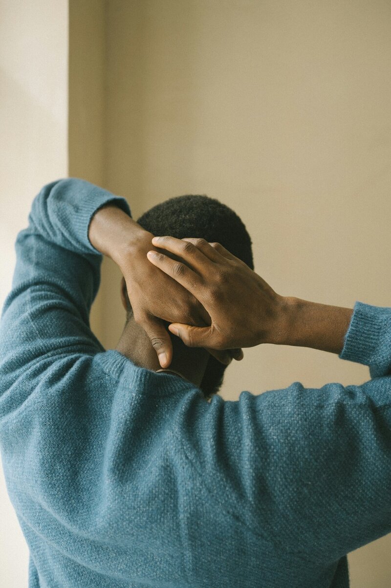 A close-up of a man in a blue sweater with his back facing the camera, resting his hands behind his head in a relaxed posture.
