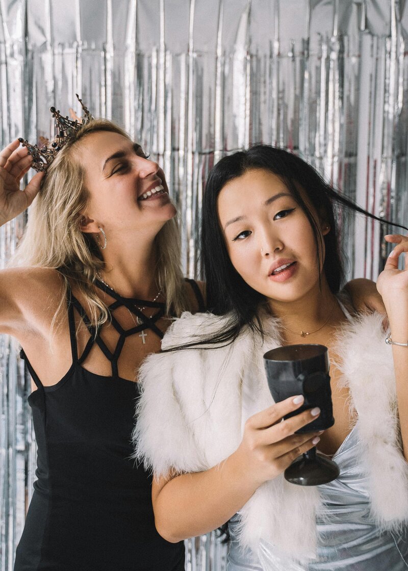 wo women celebrating at a photo booth with a metallic silver backdrop, one wearing a crown and black dress, the other in faux fur and holding a cup.