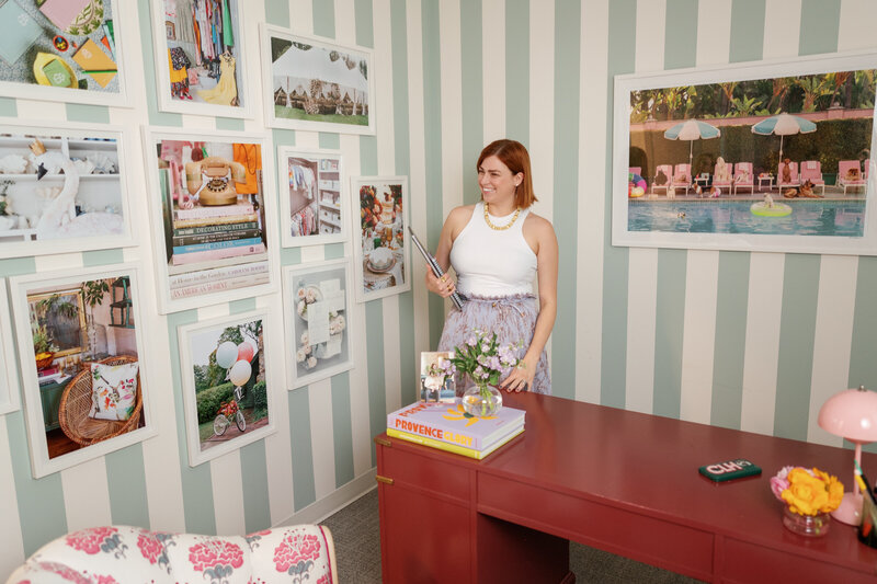 Chelsey Heslop in a white tank and purple pattern skirt holding her laptop standing next to a red desk in front of a blue striped wall with white gallery frames